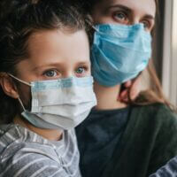 Unhappy young mother embracing upset little curly daughter with virus mask, sitting on windowsill at home, consoling sad preschool girl. Concept of coronavirus or COVID-19 pandemic disease symptoms
